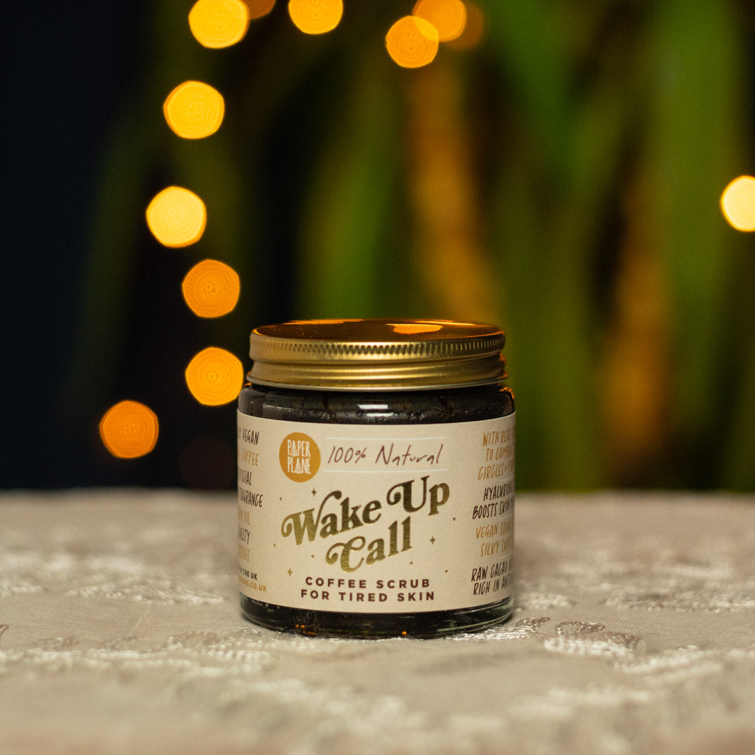 Jar of 'Wake Up Call' coffee scrub with a gold lid on a textured surface with blurred lights in the background.
