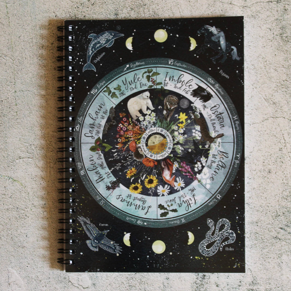 A spiral-bound notebook with a black cover featuring a detailed celestial and zodiac-themed illustration, placed on a textured light gray surface.