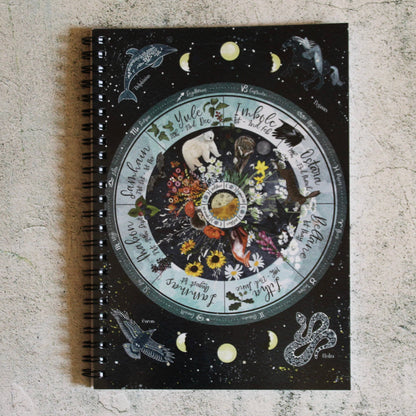 A spiral-bound notebook with a black cover featuring a detailed celestial and zodiac-themed illustration, placed on a textured light gray surface.