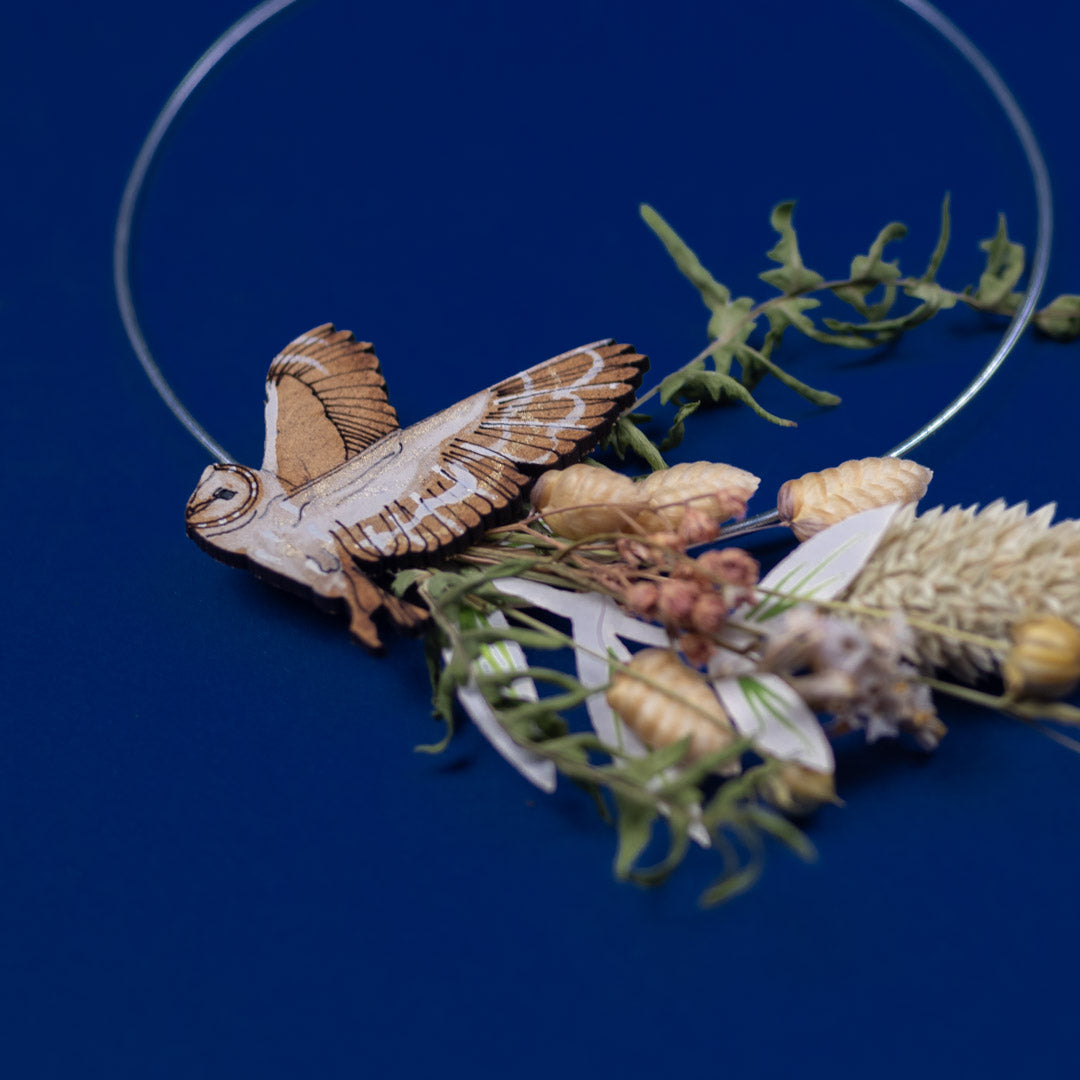 Wooden owl brooch on a blue background with dried flowers and greenery.