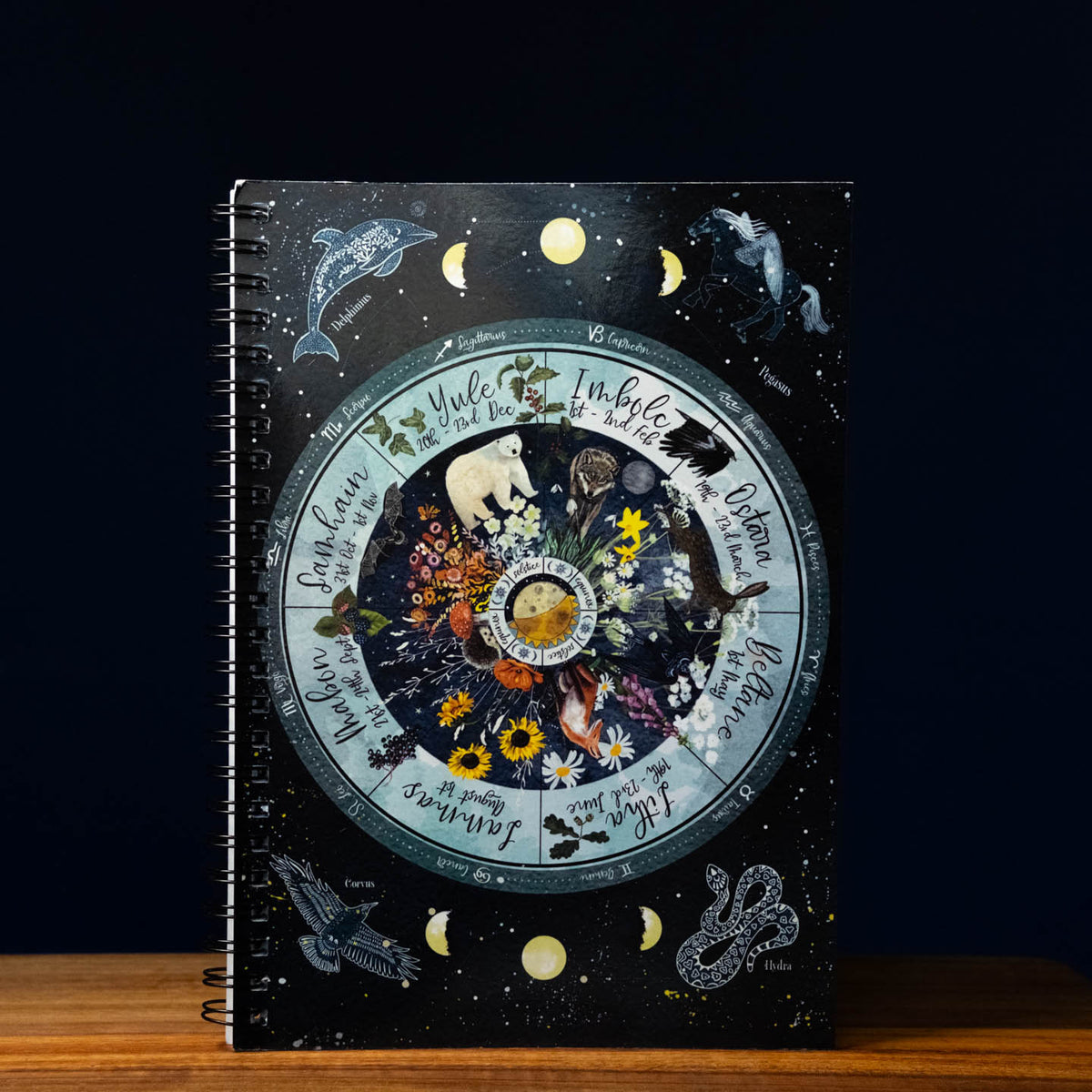 A spiral-bound notebook with a dark celestial cover featuring a large circular zodiac wheel, moon phases, stars, and illustrated animals, photographed standing on a wooden surface against a dark background.