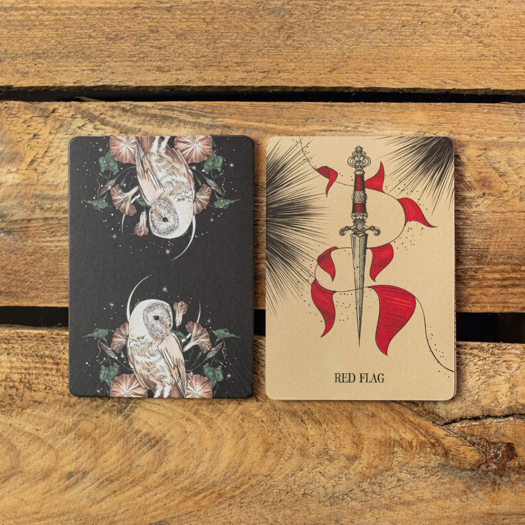 Two tarot cards on a wooden surface, one with an owl and flowers, the other with a red flag and dagger.