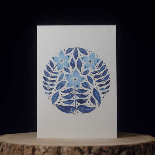 Card with blue floral design on a white background, placed on a wooden surface with a dark background.