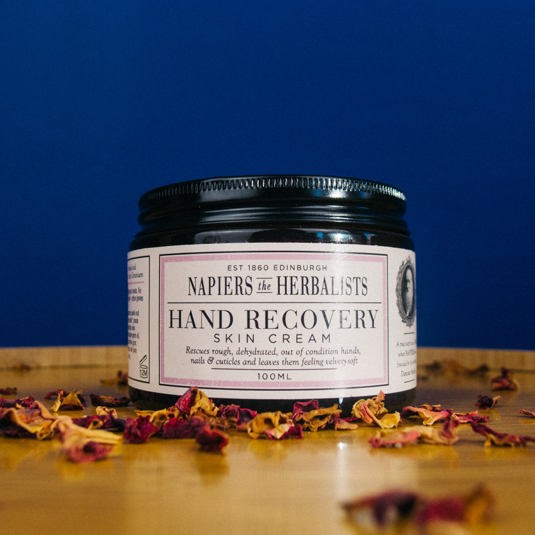 Napiers the Herbalists Hand Recovery Skin Cream jar on a wooden surface with dried flowers, against a blue background.