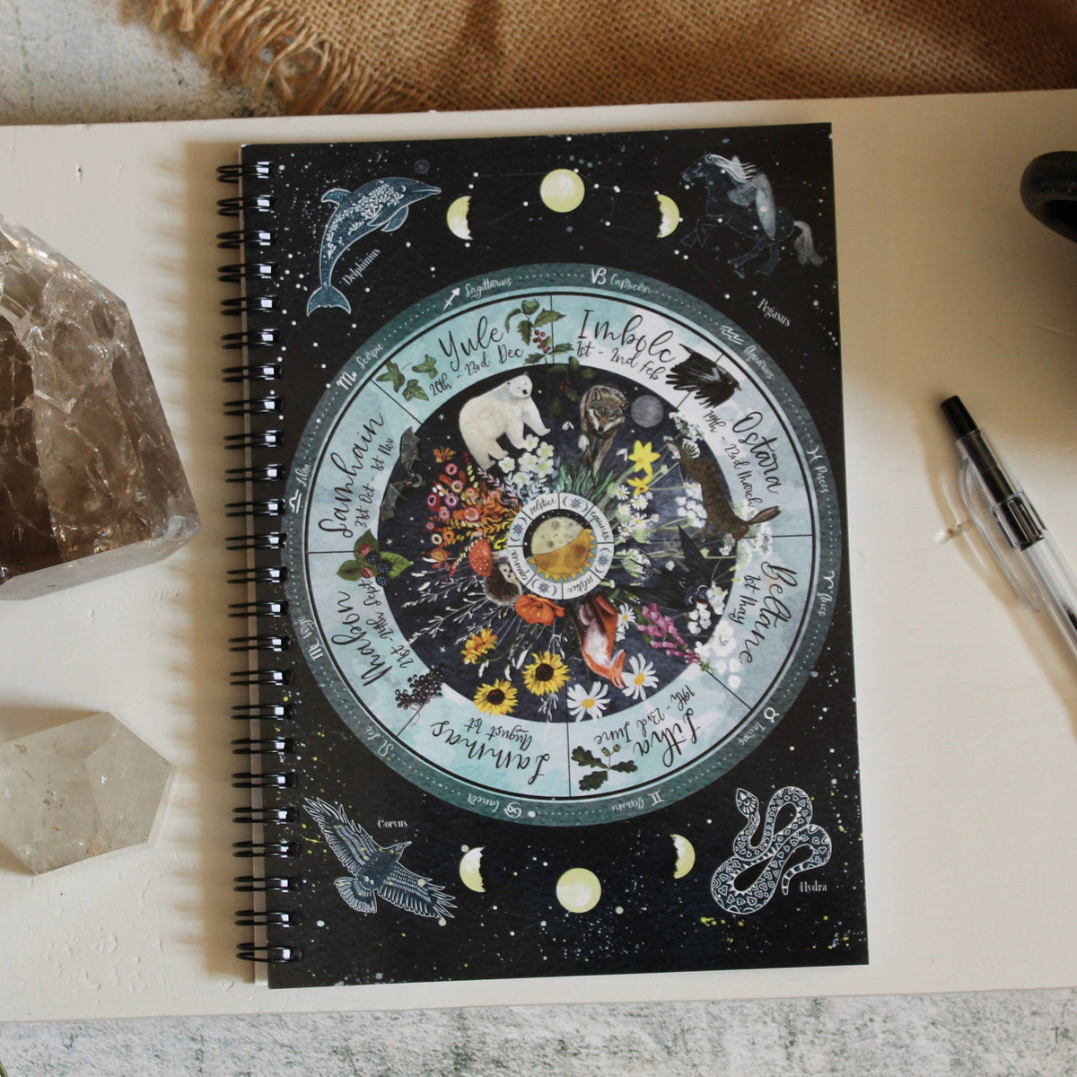 A spiral-bound notebook with a dark celestial illustration lies on a tabletop, surrounded by crystals, dried lavender, a pen, and a cup of coffee. The notebook cover features a circular astrological design with moons, stars, and floral motifs.