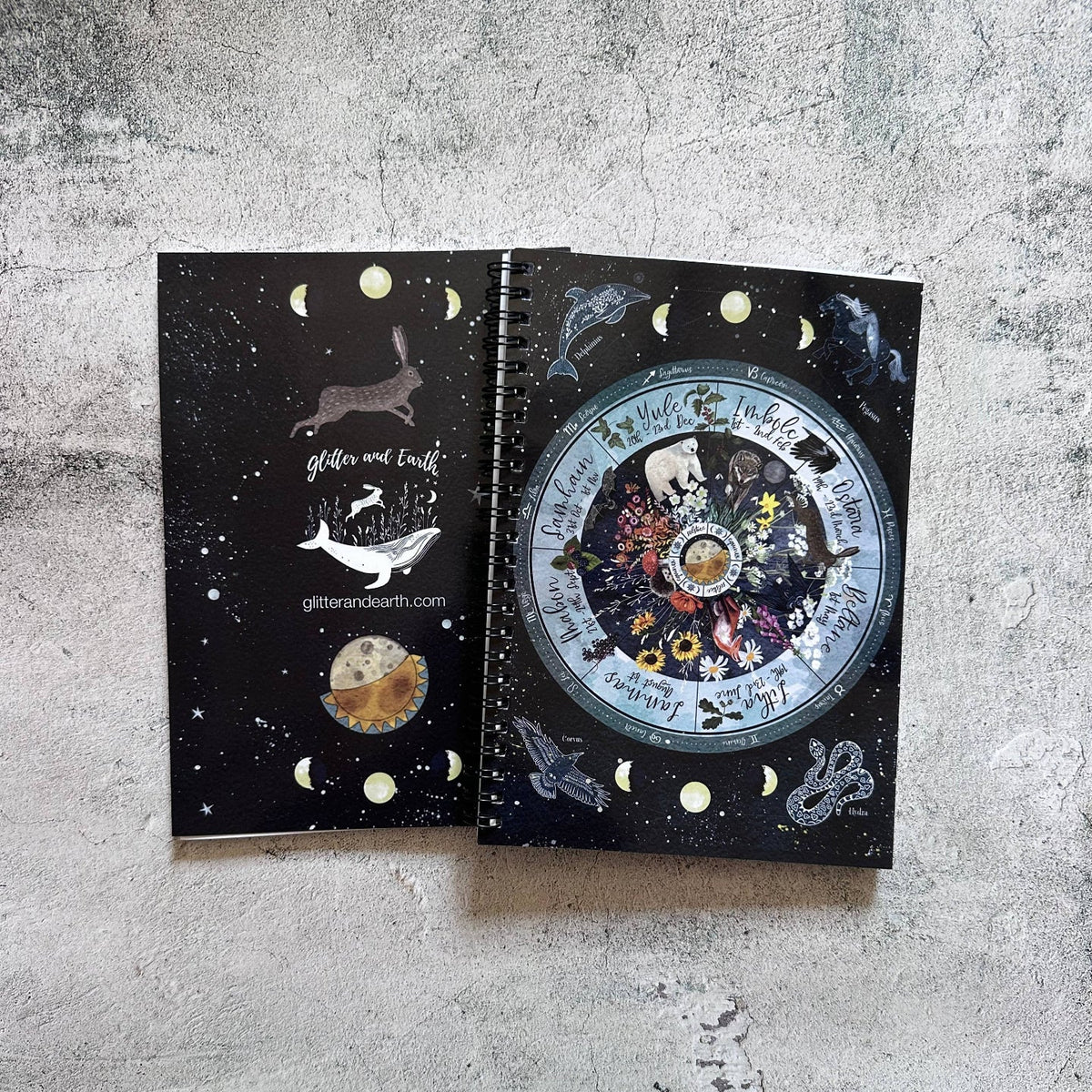 A spiral-bound notebook with a dark celestial cover design, shown open to reveal a large moon phase illustration and inner pages with moon-phase inserts, placed on a textured gray concrete surface.