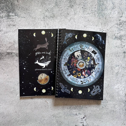 A spiral-bound notebook with a dark celestial cover design, shown open to reveal a large moon phase illustration and inner pages with moon-phase inserts, placed on a textured gray concrete surface.