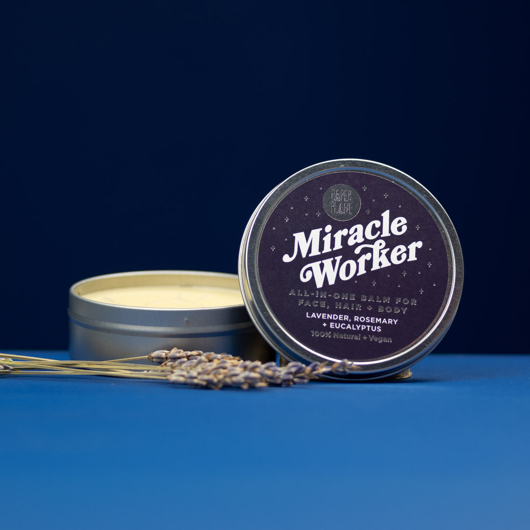 Tin of 'Miracle Worker' balm with lavender flowers on a blue background