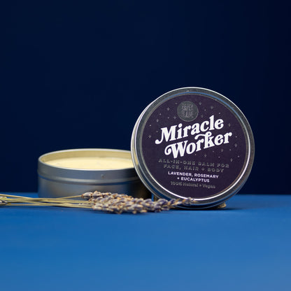 Tin of 'Miracle Worker' balm with lavender flowers on a blue background