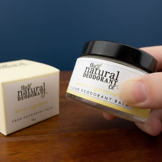 Hand holding a jar of 'The Natural Deodorant Co.' deodorant balm on a wooden surface.