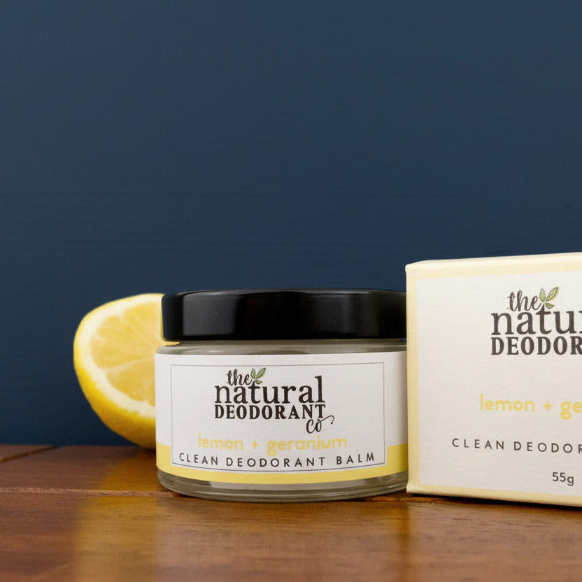 Jar of 'The Natural Deodorant Co.' lemon and geranium deodorant balm on a wooden surface with a lemon and packaging in the background.