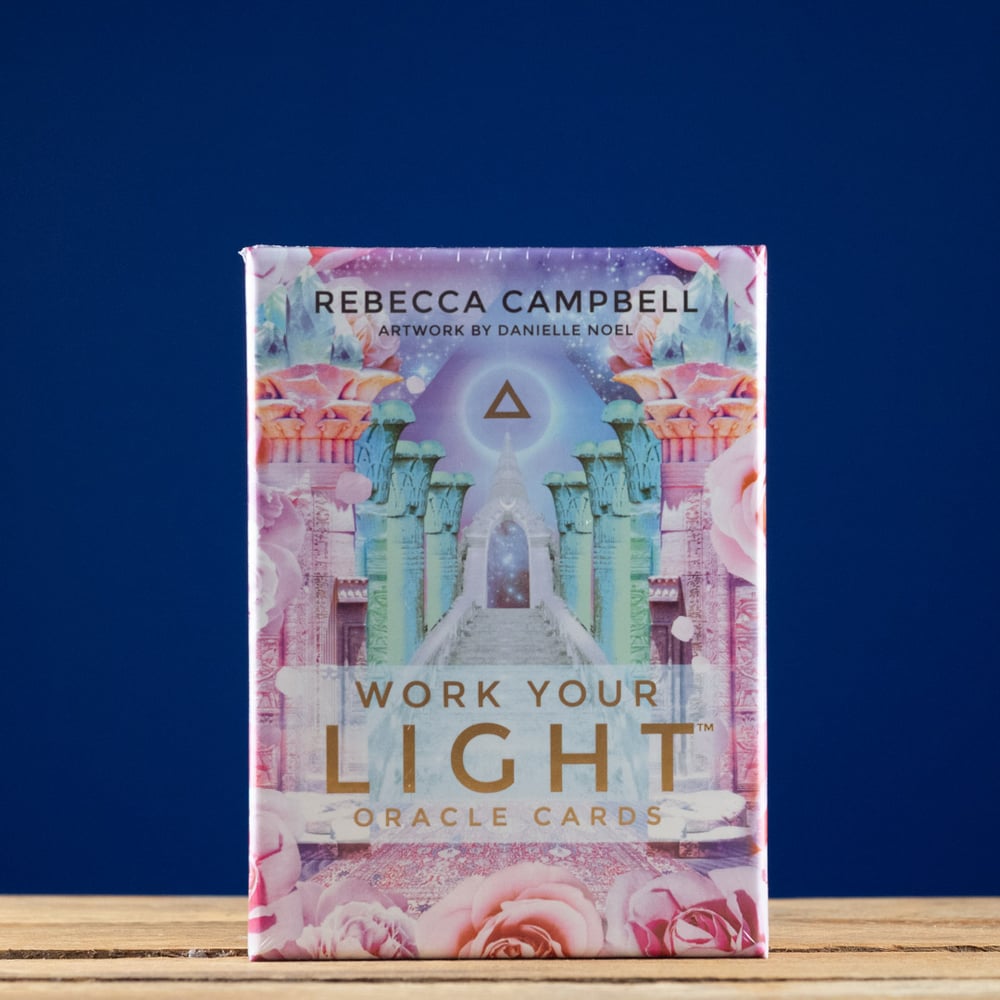Pack of 'Work Your Light' Oracle Cards by Rebecca Campbell on a wooden surface with a blue background